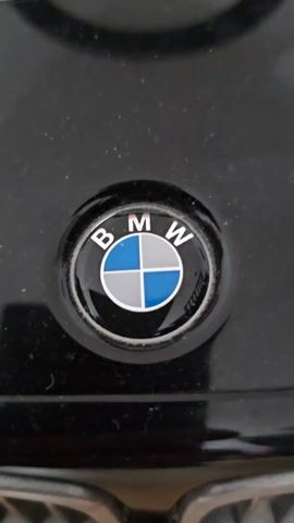 The video shows a close-up of the front of a black BMW toy car, followed by a zoom out to reveal the entire toy car. The car has a red seat, a black steering wheel, and a silver grill. The hood of the car has the words "50 JAHRE BMW M" written on it, along with the BMW logo. The car is sitting on a light brown wooden floor.