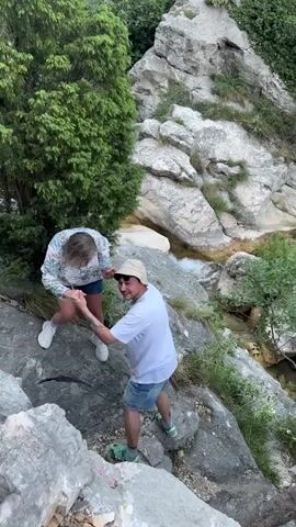 The video features two individuals, a man and a woman, navigating rocky terrain outdoors. The woman, with blonde hair, is wearing a patterned long-sleeved blouse, denim shorts, and white sneakers. She is standing on a higher rock. The man, wearing a white t-shirt, denim shorts, a light-colored bucket hat, and green/black sneakers, is positioned slightly below her. He is holding her hands, appearing to assist her or stabilize her on the uneven ground as she steps down.

They are situated on a steep, rocky incline next to a flowing stream or small river, visible in the background and at the bottom of the frame. The background consists of large, light-colored rocks and lush green foliage, including trees and bushes. The water in the stream appears clear and flows over rocks, creating small rapids and pools.

The lighting suggests it is daytime, with bright natural light illuminating the scene. The weather appears clear and pleasant. Both individuals seem focused and cooperative, indicating a shared outdoor activity such as hiking or exploring. The woman briefly looks towards the camera, acknowledging the filming. The prominent rocks, flowing water, and dense greenery strongly suggest a mountainous or hilly area with a watercourse, consistent with the natural landscapes found in Zupci, Montenegro. No visible text is present in the video.