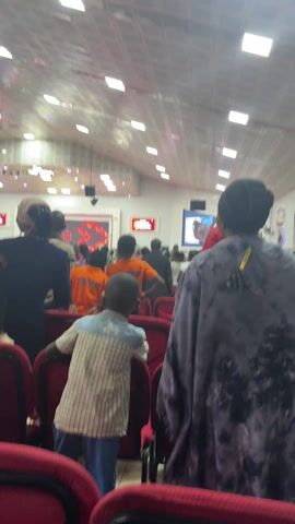 The video shows the back of a crowded room, likely a church, with people of varying ages. A few young children are in the front row, wearing orange t-shirts with text that includes "APONCREM" and "INTERNATIONAL." The room has red chairs, and there are large screens displaying images or videos at the front. The ceiling is white with a grid-like pattern of rectangular lights. People are generally standing and facing the front, seemingly engaged in an event taking place there.