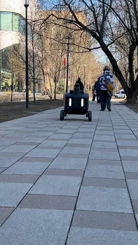 The video shows a black delivery robot with six wheels traveling down a brick-paved walkway. The robot has two vertical rectangular lights on its front, and an antenna with a small red flag. The background includes bare trees, a building with a glass facade, and people walking in the same direction as the robot. It's daytime and the sky is clear.