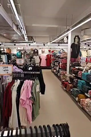 The video pans across a brightly lit clothing store. Racks of women's apparel, including dresses, blouses, and trousers, are displayed. Price tags indicating INR 299 and INR 499 are visible. A large price tag displaying INR 599 is also prominent. The store appears to be well-stocked, with multiple aisles of clothing. A large poster featuring a woman is displayed on a pillar. The ceiling is industrial, with visible conduits and lighting fixtures.
