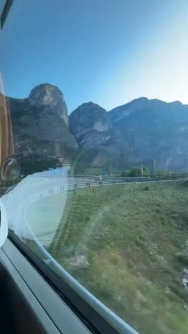 A vertical video shows a view from the window of a moving vehicle. The window reflects the interior of the vehicle, including a white headrest on the left side and a light colored frame. Outside the window, a winding road curves to the right, bordered by a metal guardrail and a grassy embankment. In the distance, a mountainous landscape under a clear blue sky is visible. Traffic signs, including a give way sign and a roundabout sign, are present near the road. The sun is low in the sky, casting a soft light on the scene.
