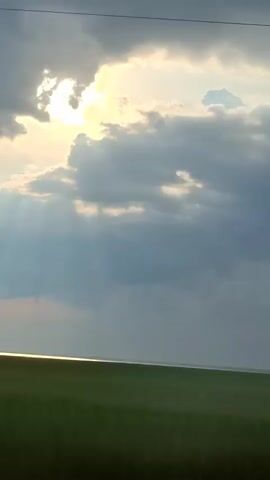 The video shows a view from a vehicle moving through a landscape. The sky is filled with dramatic clouds, with the sun breaking through in the upper left, casting visible rays. Below the sky is a flat, green expanse that appears to be a field or plain, stretching to the horizon. A bright, reflective strip runs along the horizon, suggesting the presence of water or a very smooth, flat surface. Two thin, dark lines, likely power or telephone wires, are visible in the upper portion of the frame. The footage has a slight blur, indicating motion.