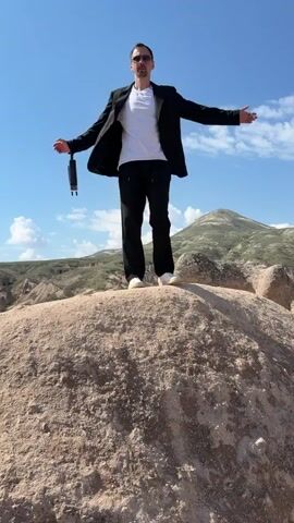 A man stands triumphantly on a large, rounded rock formation against a clear blue sky with sparse clouds. He is dressed in a white t-shirt, black blazer, black trousers, and white sneakers, holding a small black bag in his left hand. His arms are outstretched, initially looking straight ahead, then turning his head and body to his left, as if admiring the expansive view. In the background, rolling hills and distinctive rock formations, characteristic of Ürgüp, Turkey, stretch under the bright daylight. The scene suggests a feeling of freedom and appreciation for the natural landscape. There is no visible text.