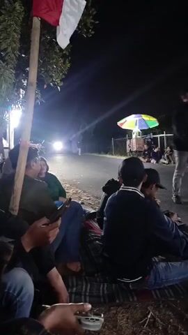 A group of people are sitting on mats on the side of a road at night. They are watching a motorcycle race. The road is lit by artificial lights. There are trees and bushes in the background. A flag is flying on a pole on the left side of the image. There is a colorful umbrella in the background. The people are dressed in casual clothing. Some of them are holding phones. The motorcycle is moving at a high speed.