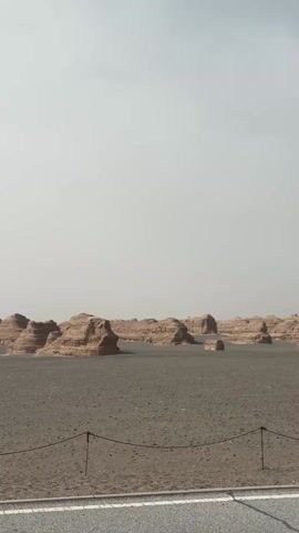 The video captures various perspectives of a vast, arid desert landscape featuring striking yardang rock formations under a hazy, overcast sky.

The initial shot at 0:00 shows a wide expanse of dusty terrain with numerous large, weathered rock formations resembling ancient structures scattered across the horizon. A dirt path, delineated by a simple rope barrier on wooden posts, extends into the distance where several people are seen walking.

At 0:01, the camera pans to reveal an asphalt road with clear white and yellow markings. The road runs alongside the desert, with more yardangs visible in the background. The sky remains muted and hazy.

The pan continues at 0:02, bringing into view a wooden information sign, partially obscured, next to the road and the desert. The unique geological formations continue to dominate the background.

At 0:03, a group of people, dressed in casual attire suitable for desert exploration, are seen walking on the dusty ground. They appear to be heading towards a prominent heart-shaped structure, possibly an art installation or photo opportunity, made of natural materials. The yardangs provide a dramatic backdrop to their activity.

The final shot at 0:04 shows a longer section of the rope-marked path, leading further into the desert towards more rock formations. Distant visitors continue to explore the unique environment.

The scene depicts a tourist attraction, likely a geopark, known for its distinctive yardang landforms. The overall impression is one of exploring a unique natural wonder in a dry, hazy environment, possibly near Dunhuang City, China, known for its Yardang National Geopark. The time of day appears to be during daylight hours, but the hazy conditions obscure direct sunlight. No clearly legible text is visible in the video.