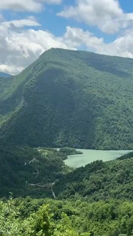 This is a scenic, elevated video of a vast, lush green mountain range with a body of water nestled in a valley. The sky above is partly cloudy with white clouds against a blue backdrop. The mountains are densely covered with trees, creating a textured, undulating landscape. A winding road is visible in the valley, disappearing into the trees. The water in the valley appears to be a light green or turquoise color. The overall impression is one of natural beauty and tranquility. The video appears to be a static shot with no visible movement or subjects.