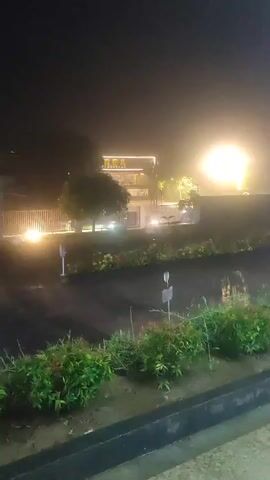 This media file shows a nighttime scene in Cotbadatunong, Indonesia. The foreground is dominated by a dark, wet surface, likely a road or parking lot, with a small row of green bushes in front of it. Beyond the bushes, several cars are parked. A white hatchback is prominently visible in the center, with its front end angled towards the viewer. To its left, another white car is partially visible, and further back, a black car is also present.

In the background, a building with illuminated signage reading "ZAK" is visible. The building appears modern with several levels and bright, yellow-orange lights. The sky is dark, indicating nighttime, and there's a hazy, atmospheric quality to the scene, possibly due to rain or humidity. Several streetlights and building lights are casting a glow, illuminating parts of the scene and creating reflections on the wet surfaces. The overall mood is somewhat serene, despite the artificial lighting. There are no people visible in the media file.