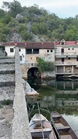 The video shows a stone bridge with stairs leading over a body of water. Two boats are docked in the water below the bridge. Buildings with red roofs and white walls are situated along the opposite bank of the water, with lush green trees and rocky hills in the background. A person is visible walking up the stairs on the bridge. The weather appears to be overcast.
