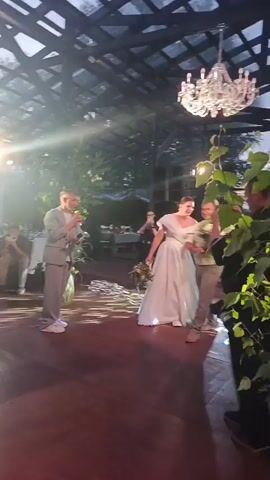 The video shows an outdoor wedding reception with a light-colored gazebo structure overhead. A person in a light-colored suit is speaking into a microphone while holding a bouquet. Another person in a wedding dress is standing nearby. A man carrying a large bouquet of flowers walks across the dance floor. Guests are seated at tables in the background. There is a large chandelier hanging from the ceiling. The lighting is bright and appears to be from natural sunlight and artificial lights.
