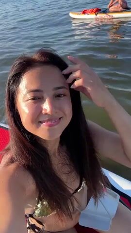 A woman with long dark hair is paddleboarding on a body of water. She is smiling and looking at the camera. She is wearing a bikini top and a light-colored sarong or cover-up. A straw hat and a paddle are visible on the paddleboard. The water is calm and the sky is blue with some clouds. In the background, there are trees and some distant boats. The video is a selfie-style shot, with the camera appearing to be held by the woman herself or attached to the paddleboard.