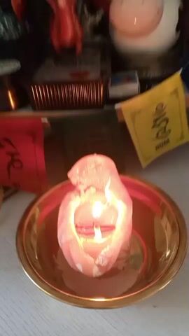 A short video shows a close-up view of a candle burning in a brass bowl. The candle is pink and appears to be melting. Behind the candle, a shelf is visible with various items, including a red dragon figurine, a white cow figurine, and several small Tibetan prayer flags. The prayer flags are colorful, with one yellow flag displaying symbols. The background includes more items on shelves, such as flowers, dried plants, and what appear to be artwork or posters on the wall. The lighting is dim, and the video is slightly blurry, giving it a soft, ethereal quality. The video appears to be taken in a home setting, possibly as part of a ritual or altar.