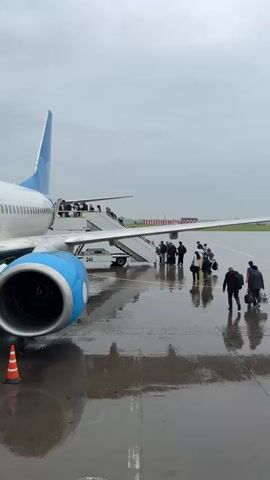The video shows a group of people boarding an airplane on a rainy day. The plane is parked on the tarmac, and a set of stairs leads up to the entrance. People are walking up the stairs, carrying luggage. The ground is wet and reflects the sky and the airplane. The sky is overcast and gray. The plane's engines are visible, one of which is in the foreground. The scene appears to be at an airport.