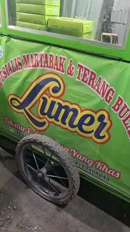 The video shows a close-up of a food cart with a green banner that reads "SPESIALIS MARTABAK & TERANG BULAN LUMER" in large lettering. Below the brand name, smaller text says "Tekstur & Rasa Yang Khas" followed by a phone number. A wheel of the food cart is visible in the foreground. In the background, there are stacks of yellow boxes, possibly containing food items. The overall scene appears to be outdoors, possibly at night, with dim lighting. There are no people clearly visible in the frame.