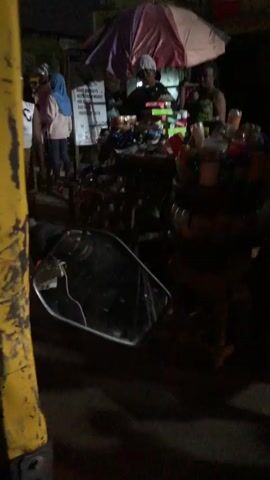 A dimly lit outdoor scene at night captures a street vendor's stall. Several people are present, including two women standing near the stall and at least two more behind it. One woman in the foreground on the left wears a white off-the-shoulder top and a patterned skirt. Another woman, in the background, is wearing a blue headscarf and white top. The stall itself is laden with numerous items, including stacks of colorful boxes, bottles, containers, and various food or drink items. A large, round umbrella, partially illuminated, is positioned above the stall. In the foreground, a reflection of a person's arm and torso is visible in what appears to be a motorcycle's side mirror. The ground is dark and appears to be paved. The overall impression is of a busy market or roadside stall operating in the evening.
