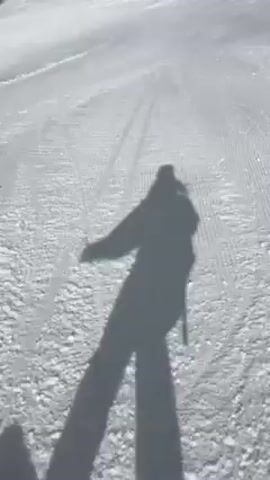 The video shows a person skiing downhill. The camera captures a first-person point of view, focused on the snow ahead and the skier's shadow. The snow appears to be well-groomed and corduroyed. The landscape includes trees on the surrounding slopes. The weather appears to be sunny. The person is skiing straight ahead.