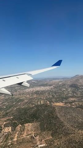 The video shows an aerial view from an airplane window. The airplane's wing, white with a blue tip, is visible in the foreground against a clear blue sky. Below, the landscape is a mix of greenery, fields, and scattered buildings forming a small town. In the distance, there are mountains and a hazy horizon. The camera is static, providing a stable view of the scene as the plane appears to be in descent. There are no visible people in the frame.