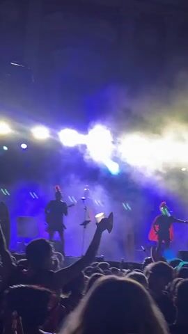 The video captures a concert performance with a band on stage and a crowd in the foreground. The stage is illuminated by bright blue and white spotlights, creating a dramatic atmosphere with visible smoke or haze. Two figures, presumably band members, are silhouetted on stage, one holding what appears to be a microphone or instrument and the other holding a guitar. The crowd is dense and dark, with many people visible from the back, some with their arms raised. The overall impression is one of an energetic live music event.