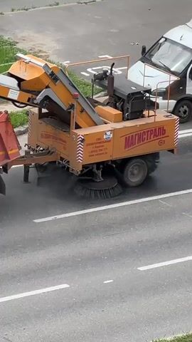 This media file captures a street sweeper truck in motion on an asphalt road, likely in Tolyatti, Russia, given the context. The truck is a bright orange and yellow industrial vehicle with multiple rotating brushes at its base, actively sweeping the road surface. Dust or debris is visibly being kicked up by the brushes.

The truck has a prominent logo that reads "МАГИСТРАЛЬ" in large, bold, white Cyrillic letters, suggesting it's part of a road maintenance or transportation company. Below this, there's smaller text which appears to be contact information and possibly a company name, including a phone number starting with "+7 (843) 294-171" and a website address.

A white van is partially visible in the background, to the right of the street sweeper, indicating it is operating on a public road. The surrounding area shows a strip of green grass and a sidewalk or paved area adjacent to the road, with some buildings or structures further back, though they are out of focus.

The lighting suggests it is daytime, possibly late morning or afternoon, with overcast conditions as there are no harsh shadows. The overall scene is functional and industrial, focusing on the task of road cleaning. There are no people visible in the frame. The motion of the brushes and the dust indicate the street sweeper is actively engaged in its work.