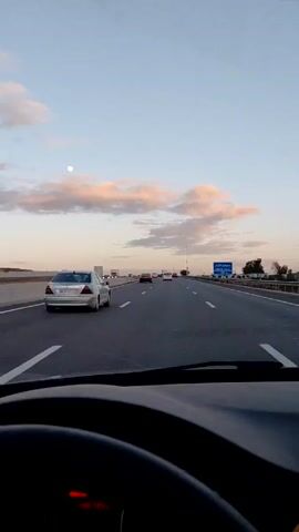 The view from inside a moving car shows a multi-lane highway with traffic. The dashboard and steering wheel of the car are visible. The road is marked with white lines. Other cars are on the highway ahead. A blue sign with white text, likely indicating a location, is visible to the right. The sky is blue with light pink clouds. A small, bright, round object, likely the moon or sun, is high in the sky. The overall impression is of a journey on a highway at dusk or dawn.