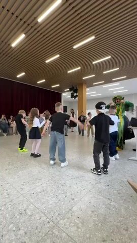 In a brightly lit hall with a suspended slat ceiling, a group of about ten children of various heights are standing in a loose circle and facing outward. They are dressed in casual clothing, including jeans, t-shirts, dresses, and sneakers. Most have their backs to the camera. In the center of the circle, an adult female in a black dress is leading the children in a dance or exercise routine. To her right, a large, green-costumed character with an oversized head is also participating.

Behind the group, a burgundy curtain is drawn along one wall, and a scattering of adults are seated watching the activity. There is a speaker on a stand beside the green figure, and a disco ball hangs from the ceiling in the middle of the hall. The hall has a speckled, terrazzo-style floor. The camera is held by someone seated, and part of a pair of denim-clad legs and a beige heeled shoe are visible in the lower right corner of the frame. The atmosphere appears to be casual and cheerful.