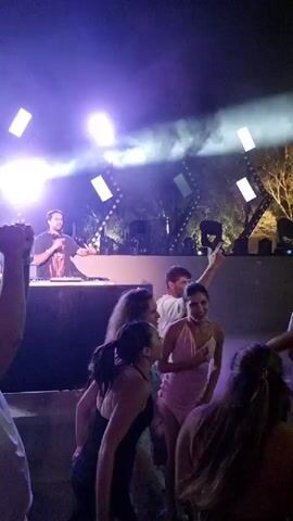 A group of people are dancing at a nighttime outdoor event. A DJ is visible behind a table with a laptop and equipment. Bright stage lights, including spotlights and geometric light fixtures, illuminate the scene. Several individuals in the foreground are dancing, some with their arms raised. The atmosphere appears to be a lively party or club setting.