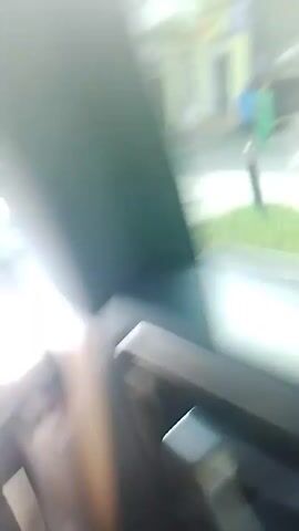 The video shows a view from inside a vehicle, looking out through the passenger window. The scene is a street with buildings on the right side of the road. The buildings appear to be shops and residential properties. There are trees and greenery visible along the street. A traffic sign that reads "no parking" is also visible. People are seen walking on the sidewalk. The sun is shining brightly, causing lens flare in the video. The camera appears to be in motion, as the scenery outside is passing by.