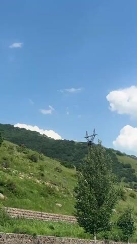 A vertical, wide shot captures a vast, clear blue sky dotted with fluffy white clouds. Below the sky, a lush green, tree-covered hillside slopes downwards. In the foreground, a tall green tree stands prominently, obscuring part of the view. To the left of the tree, a power line tower with wires is visible. In the distance, on the lower left side of the frame, a person in dark shorts and a blue top stands near what appears to be a fence or barrier. The overall scene suggests an outdoor natural environment on a sunny day.
