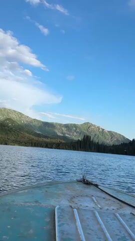This media file captures a serene view from a boat on a calm lake, likely in Arkit, Kyrgyzstan. The foreground is dominated by the weathered, blue-painted bow of the boat, with a metal mooring cleat and a dark rope visible. The lake's surface is gently rippling, reflecting the bright blue sky above, which is dotted with a few wispy white clouds.

In the mid-ground, a dense, dark green forest lines the shore, primarily composed of tall, slender evergreen trees. This forest extends to meet the foothills of imposing mountains. These mountains, with their rugged, rocky slopes and patches of green vegetation, form the backdrop of the scene. The sunlight suggests it is likely late afternoon or early evening, casting a soft glow on the landscape. The overall atmosphere is one of tranquility and natural beauty. No visible text is present.