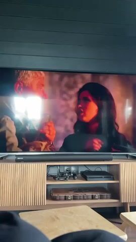 A television screen displays a scene from a film. On the left side of the screen is an older man with gray hair and a suit. On the right side of the screen is a young woman with dark hair, wearing a dark jacket. The woman is holding a small object to her ear. The television is mounted above a wooden entertainment unit. The entertainment unit has a soundbar below the television, and shelves with various electronic devices and a small suitcase. In the foreground, a wooden table is visible. The lighting suggests an indoor setting. The overall impression is of someone watching television.
