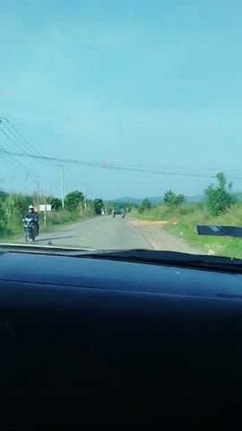 The video shows a car driving down a rural road on a clear day. There are power lines running along the left side of the road. Several people on motorbikes are traveling ahead. Trees and other greenery line both sides of the road. The dashboard of the car is visible in the foreground.