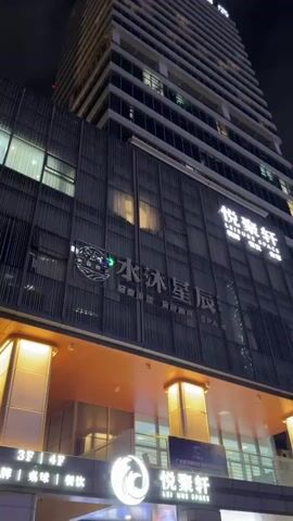 The video showcases a modern, multi-story building at night, likely a hotel or a commercial establishment, in Huangpu District, China. The building features a sleek, contemporary design with extensive glass windows and metallic cladding. The overall atmosphere is one of urban sophistication, with the building illuminated by artificial lights that create a warm glow from within and accent the architectural features.

The camera pans upwards, emphasizing the height and grandeur of the structure against the dark night sky. The lower levels of the building are particularly well-lit, revealing an entrance area with large windows and prominent signage. The lighting from below casts a golden hue onto the underside of an overhang, creating a welcoming and somewhat dramatic effect.

Visible text includes Chinese characters and English words, suggesting the names of businesses or services housed within the building. One prominent sign displays "水沐星辰" in Chinese characters, which translates to "Water Bathes the Stars," and the English tagline "LEISURESPACE." Another sign lower down also shows "LEISURESPACE." There are also indications of floors, such as "3F 4F". The overall impression is of a well-maintained, upscale establishment operating after dark. The lighting and the building's architecture convey a sense of modernity and perhaps a hint of luxury. No people are clearly visible in the frames. The weather appears to be clear, as indicated by the dark, cloudless sky.