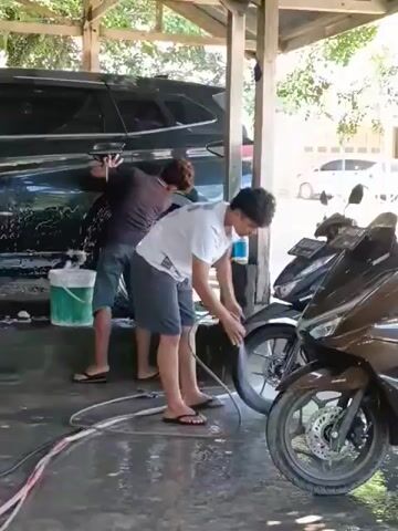 Two individuals are washing vehicles under an open-air shelter. The person on the left, wearing a dark t-shirt and shorts, is standing next to a dark-colored car and appears to be washing it with a green bucket. The person on the right, wearing a white t-shirt and grey shorts, is focused on washing the front wheel of a black motorcycle with a hose. Both individuals are wearing flip-flops. The ground is wet and soapy. There are other motorcycles parked nearby. The environment suggests an outdoor car wash or a similar service area.