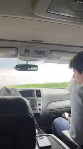 The interior of a car is shown in the video. A young man with curly brown hair is sitting in the passenger seat, looking down and to the right. He is wearing a gray hoodie and blue jeans. His hands are in his lap. The dashboard and steering wheel are visible in the foreground. The windshield shows a green field and a cloudy sky. A black smartphone is on the center console. Music is playing. A rearview mirror is visible. The car appears to be a Nissan.
