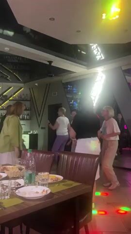 This video captures a lively scene inside a dimly lit establishment, likely a restaurant or bar, in Orsha, Belarus. Several people are present, with a group of women appearing to dance or socialize in the center of the frame. They are dressed in casual attire, with one woman in a green jacket, another in a white t-shirt and purple pants, and others in darker tops and white or beige bottoms. In the background, a bar area is visible with bottles on shelves, suggesting a place that serves drinks. The ceiling features modern, angular designs and reflective surfaces, illuminated by vibrant, colored lights that cast colorful glows on the floor and walls, creating a festive atmosphere. The time of day appears to be evening or night, given the interior lighting. In the foreground, a table is set with plates, glasses, and food, including what looks like French fries and pizza, indicating a dining experience. The overall mood seems to be one of enjoyment and celebration.