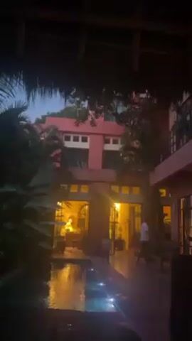 The video shows a courtyard scene at dusk or dawn.  In the foreground, a thatched roof partially obscures the view.  A small, dark pool of water reflects the lights from the building behind. Tropical plants are visible to the left of the pool. The main building is painted a muted pink color, with multiple levels and windows. Two open doorways are visible on the ground floor, revealing a warmly lit interior with furniture. A child walks across the illuminated area in front of the doorways, heading right.  A person wearing a white shirt stands in the right corner, looking away from the camera.