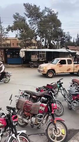 The scene captures a public space on what appears to be an overcast day. Several motorcycles are parked close to the camera. A tan pickup truck with a camouflage pattern drives across the frame. At least two individuals are visible in the back of the truck. One of them is operating what looks like a heavy weapon mounted in the truck bed. People are seen walking on the street, appearing to pay little attention to the truck. Buildings and trees form the backdrop of the scene. The general environment suggests a location that is potentially experiencing conflict or instability.