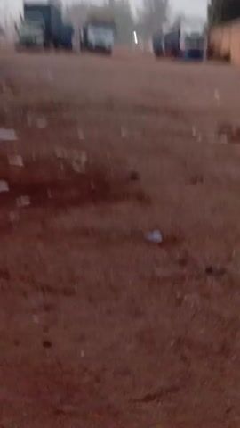 The camera is held at ground level, pointing towards a dusty, reddish-brown, dirt road with scattered litter. In the background, several large trucks are parked on the side of the road, obscured by a hazy or foggy atmosphere. A low wall runs along the right side of the frame, with glimpses of a building behind it. A person’s legs briefly come into view in the foreground. The camera is shaky.