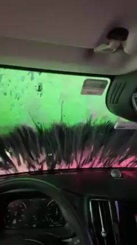 The video shows the interior of a car going through an automatic car wash. The view is from the driver's seat, looking through the windshield as large, wet brushes scrub the glass. The car wash is illuminated with pink and green lights, creating a colorful spectacle on the windshield.