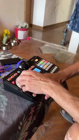 A person is seen operating a DJ controller. The controller is black and has various buttons and a turntable. The person's hands are visible, actively engaged with the controls. The background appears to be an indoor environment with wooden floors and some construction or renovation materials visible, including a red bucket and tools. A phone is also on the table.