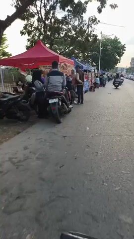 A low-angle, first-person perspective video shows a street market at dusk. Several food stalls with colorful awnings are set up along the side of a paved road. A few motorcycles are parked near the stalls, and people are seen walking and interacting. The overall scene suggests a lively evening marketplace.