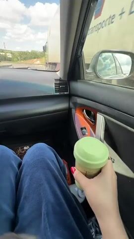 The video shows a person sitting in the passenger seat of a car, holding and shaking a light green shaker bottle with red nail polish on their nails. The bottle has measurement markings on the side. The person is wearing blue pants. Through the car window, a highway is visible with a large truck and a cloudy sky. The car's interior features a black dashboard and woodgrain accents on the door.