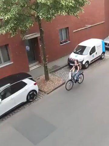 The video shows a street scene. A white van with blue accents and a logo is parked on the right side of the street. A grey SUV is parked behind the van. A white car with a black roof is parked on the left side of the street. A person is riding a bicycle in the middle of the street, moving from left to right. The person is wearing jeans and a light-colored top and has a backpack. There are trees with green foliage along the sidewalk. A brick building is visible on the right side of the street. The street surface is paved with cobblestones and asphalt. The lighting suggests it is daytime.