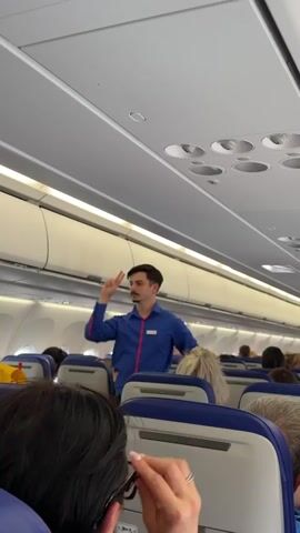 The video captures the interior of an airplane cabin, focused on a male flight attendant giving instructions to passengers. The flight attendant, dressed in a blue button-down shirt, stands in the aisle with his back to the camera, demonstrating safety procedures with arm gestures. He has dark hair and a neatly groomed mustache. The cabin is filled with passengers seated in blue cushioned seats. Overheard luggage compartments run along the sides of the cabin. The lighting inside the plane is bright. The overall atmosphere suggests the pre-flight safety demonstration on an airplane.