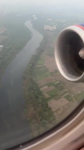This is an aerial view from an airplane window looking down at a landscape. In the foreground, part of an airplane engine is visible on the right side of the frame. Below, a river winds through the landscape, bordered by trees and agricultural fields. Buildings and what appear to be apartment complexes are scattered throughout the scene, with a general haze obscuring the distant horizon. The lighting suggests it could be daytime, possibly early morning or late afternoon, given the soft light and the presence of a slight atmospheric haze.