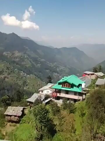 The video shows a panoramic view of a mountainous region with lush green forests covering the slopes. Several houses, some with distinctive green roofs, are nestled on the hillsides. A few smaller, rustic structures with stone roofs are also visible. The sky is blue with some scattered white clouds. The camera pans across the landscape, showcasing the natural beauty and human habitation in the area.