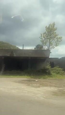 This is a short video clip with a blurry, shaky perspective, likely from a moving vehicle. The footage captures scenes of rural or semi-rural environments.

The first segment shows a weathered, concrete structure with an open, covered area underneath. It appears to be an abandoned or disused building, with some overgrowth. The sky is overcast with heavy clouds.

The video then transitions to a view of trees and foliage, creating a sense of passing through a natural landscape. Another segment shows a similar weathered building, again suggesting abandonment or decay.

A later part of the video reveals a dirt road leading into a more wooded area, with some scattered houses and a tall, dark, cylindrical structure that resembles a tower or chimney, heavily overgrown with vegetation. The sky here is a mix of clouds and patches of brighter light, suggesting a transition in the weather or time of day. A stone fence is visible in the foreground.

The overall impression is one of traversing through a somewhat neglected or natural landscape, with glimpses of old structures. There is no visible text in the video. The activity is simply the act of moving through these environments. The weather appears to be cloudy or overcast for much of the clip. The location cues suggest a remote or less developed area, consistent with the provided location context of Bzypta, Abkhazia.