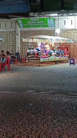 THE VIDEO SHOWS A SMALL CAROUSEL RIDE AT NIGHT. THE CAROUSEL IS ILLUMINATED WITH BRIGHT, MULTICOLORED LIGHTS AND FEATURES SEVERAL SMALL VEHICLE-SHAPED SEATS, INCLUDING CARS AND MOTORCYCLES. THE RIDE IS SET UP ON A PAVED SURFACE, POSSIBLY A STREET OR SIDEWALK, IN FRONT OF A BUILDING WITH A LARGE SIGN THAT READS "TOKO AZIFA JAYA." THERE ARE A FEW PEOPLE VISIBLE IN THE FOREGROUND, SITTING ON RED PLASTIC CHAIRS. A FEW MOTORCYCLES ARE PARKED NEARBY. THE OVERALL ATMOSPHERE IS THAT OF A NIGHT MARKET OR FAIRGROUND.