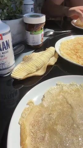 A close-up shot of crepes on white plates sits on a dark reflective table. A jar of Nutella, a spoon, and a knife with butter are visible on the table. In the background, a person is picking at a crepe. The scene then shifts to a view of a wooden deck and a wooden fence under a blue sky with white clouds. A tiled roof of a building is visible. The camera then pans up to the sky.