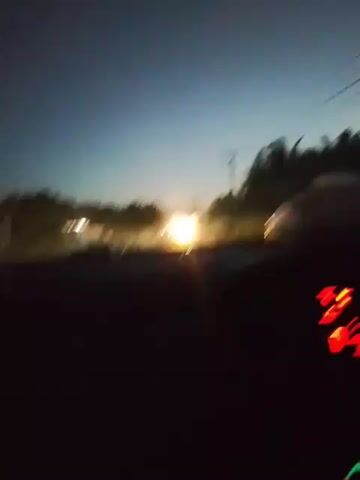 A first-person perspective video shows a car driving at dusk on a road. The sky is a deep blue with some lighter hues near the horizon, suggesting the sun has recently set or is about to rise. Trees and power lines are visible along the side of the road, with some scattered lights from houses or businesses. The car's dashboard is illuminated with red digital displays, showing information that is not clearly decipherable. The road ahead appears to be paved and leads into a faintly lit area. There are no people or other vehicles clearly visible in the frame.